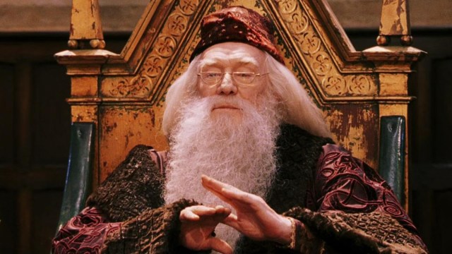 A wizened headmaster for a school of wizards, Dumbledore, gestures with his hands as if to calm a crowd while his long white hair and beard make the impression of being very wise. He also sports a patterned cap and glasses as he sits on a golden chair fit for a person of authority.