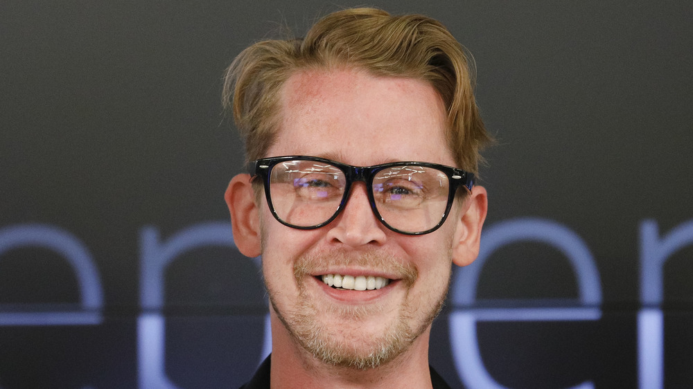 Macaulay Culkin, wearing black glasses, smiles for the camera. 