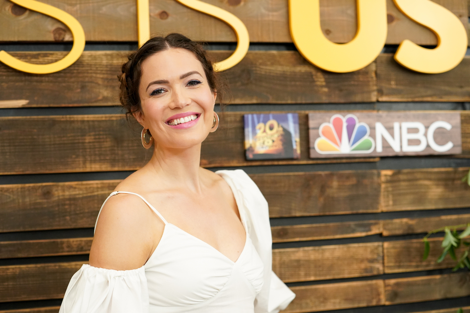 Actress Mandy Moore wears a white top and stands in front of a wooden backdrop with signage for NBC, 20th Century Fox, and the TV show This is Us.
