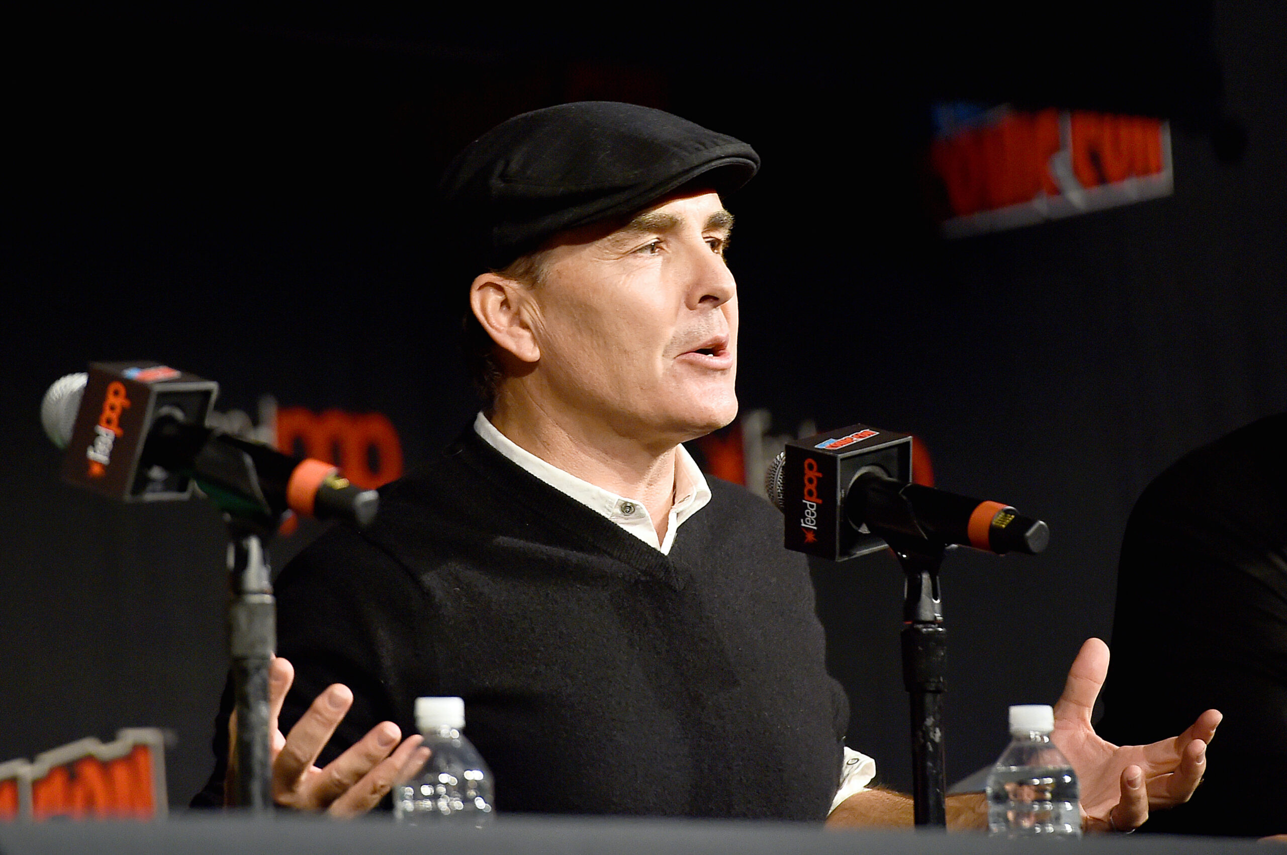 Nolan North (a white man in his late forties) wearing a flat cap and talking on a panel