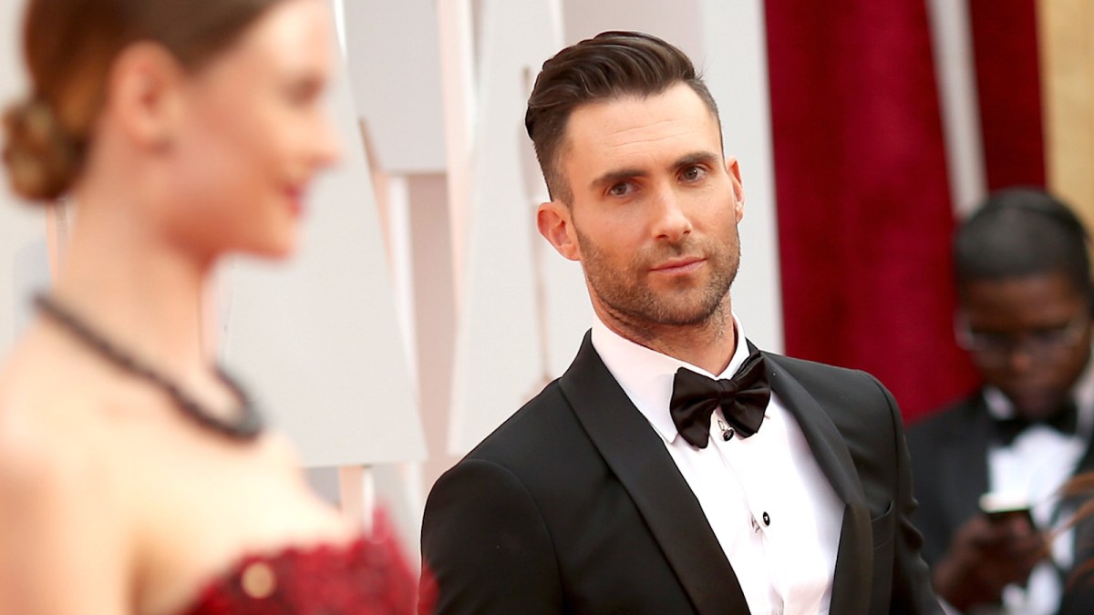 Adam Levine dressed in a black and white suit staring at the blurred profile of his wife, Behati Prinsloo