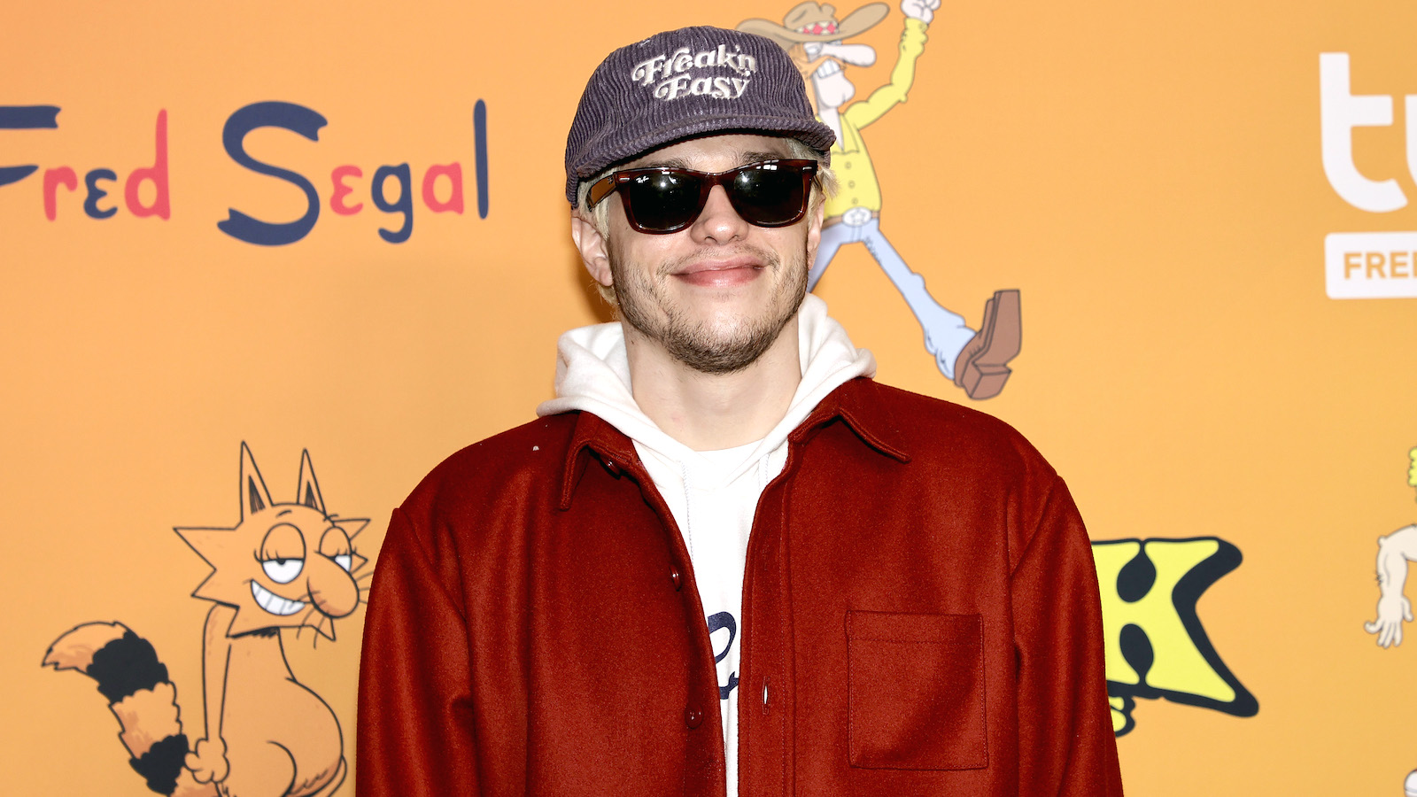 Pete Davidson is wearing sunglasses and a hat that says "Freak'n Easy" on a red carpet. 