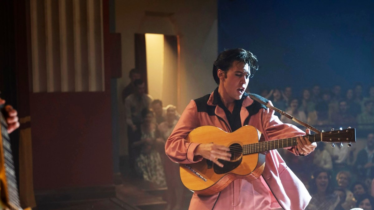 Austin Butler performing as Elvis Presley, wearing a pink and black suit while singing and playing a guitar on stage