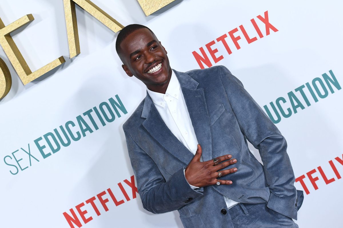 Ncuti Gatwa in a blue velvet suit jacket and white shirt, smiling.