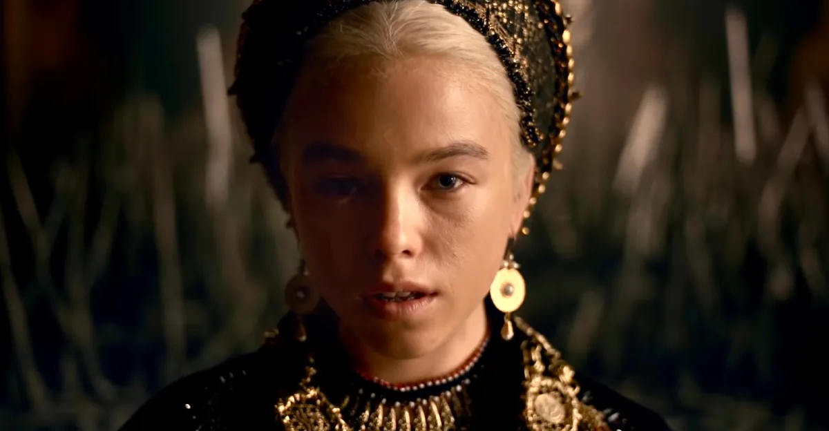 Milly Alcock as young Princess Rhaenyra Targaryen with ornate headdress, jewelry and clothing, faces the viewer head--on, the camera framing a tight shot of her face