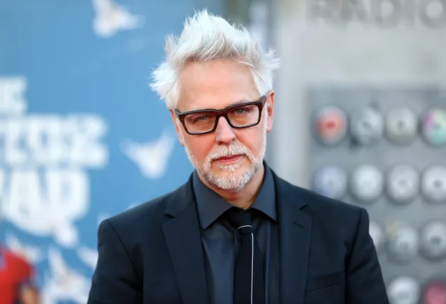 James Gunn (a white man with white hair and a white beard) looking straight at the camera and wearing a dark suit.