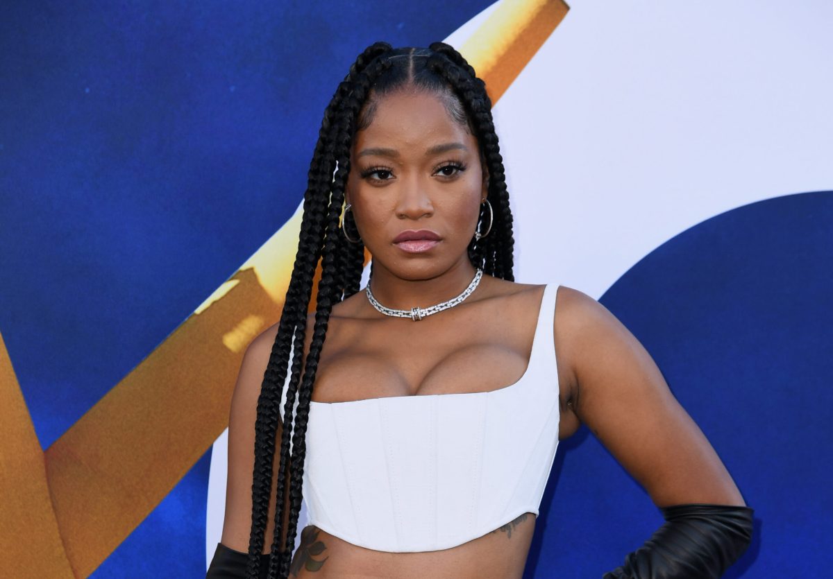 Keke Palmer (a Black woman wth long braids) with a white crop top on, posing with a hand on her hip looking straight at the camera.