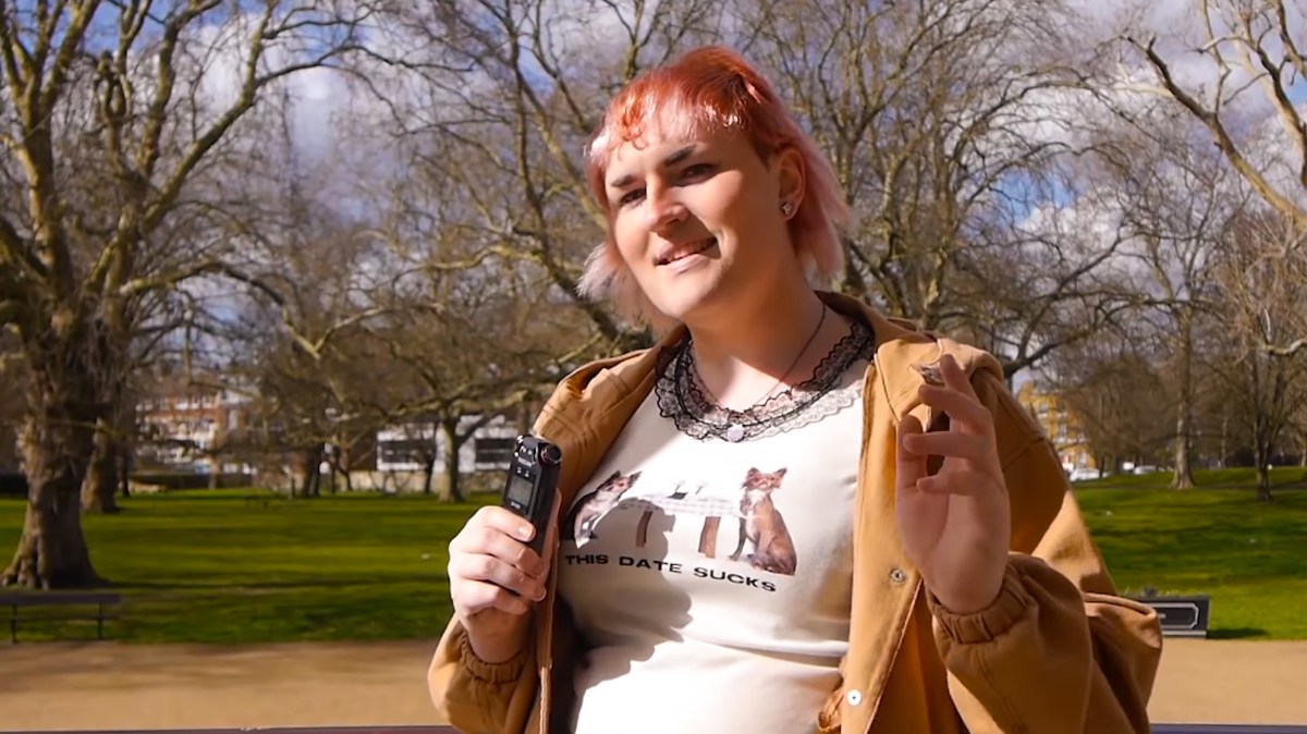 YouTuber Sophie From Mars is pictured outdoors, wearing a t-shirt and tan jacket and a colorful pink and orange hairdo with bangs, smiling and speaking into a microphone while looking into the camera