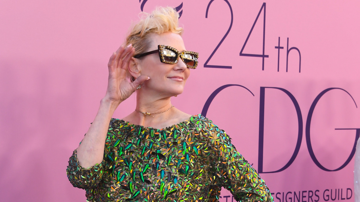Anne Heche at a premiere stands in a green dress against a pink background, wearing sunglasses and cupping her hand against her ear