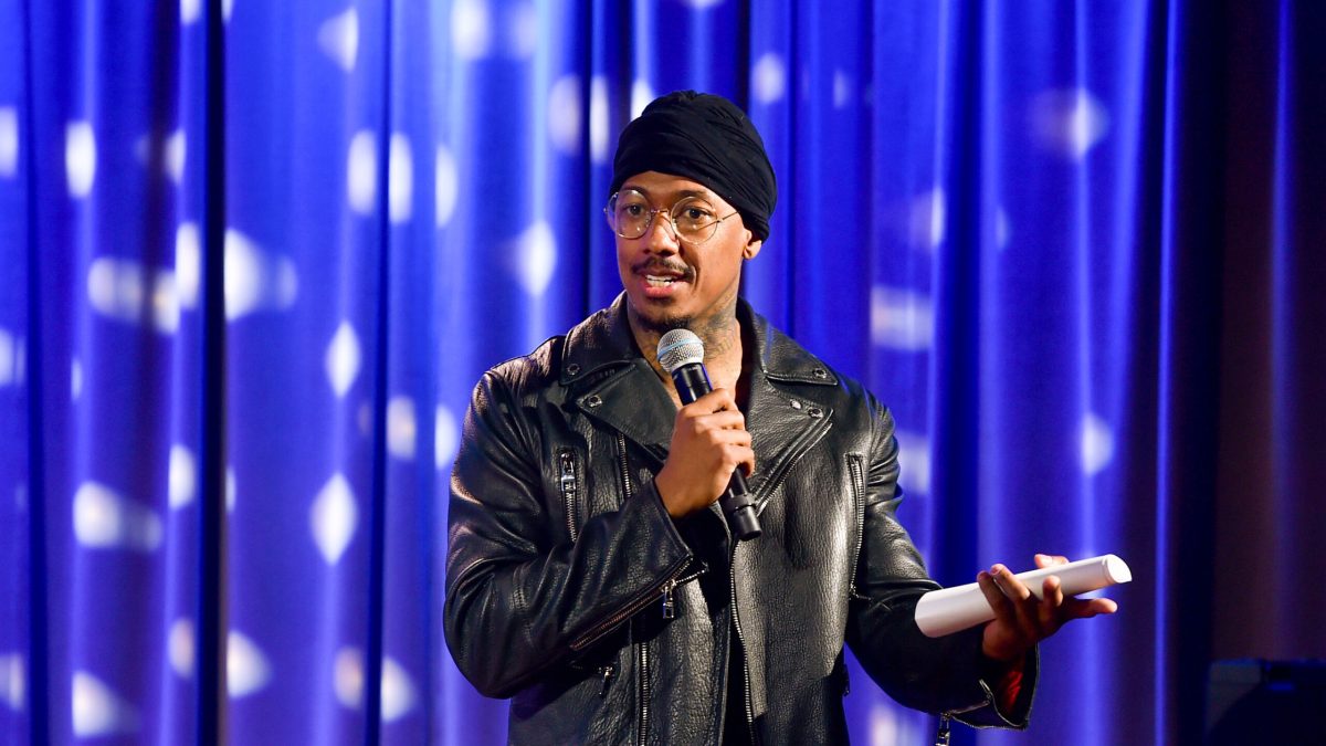 Nick Cannon is shown from the waist up, wearing a black motorcycle jacket, wire rimmed glasses and a black beanie, speaking into a microphone