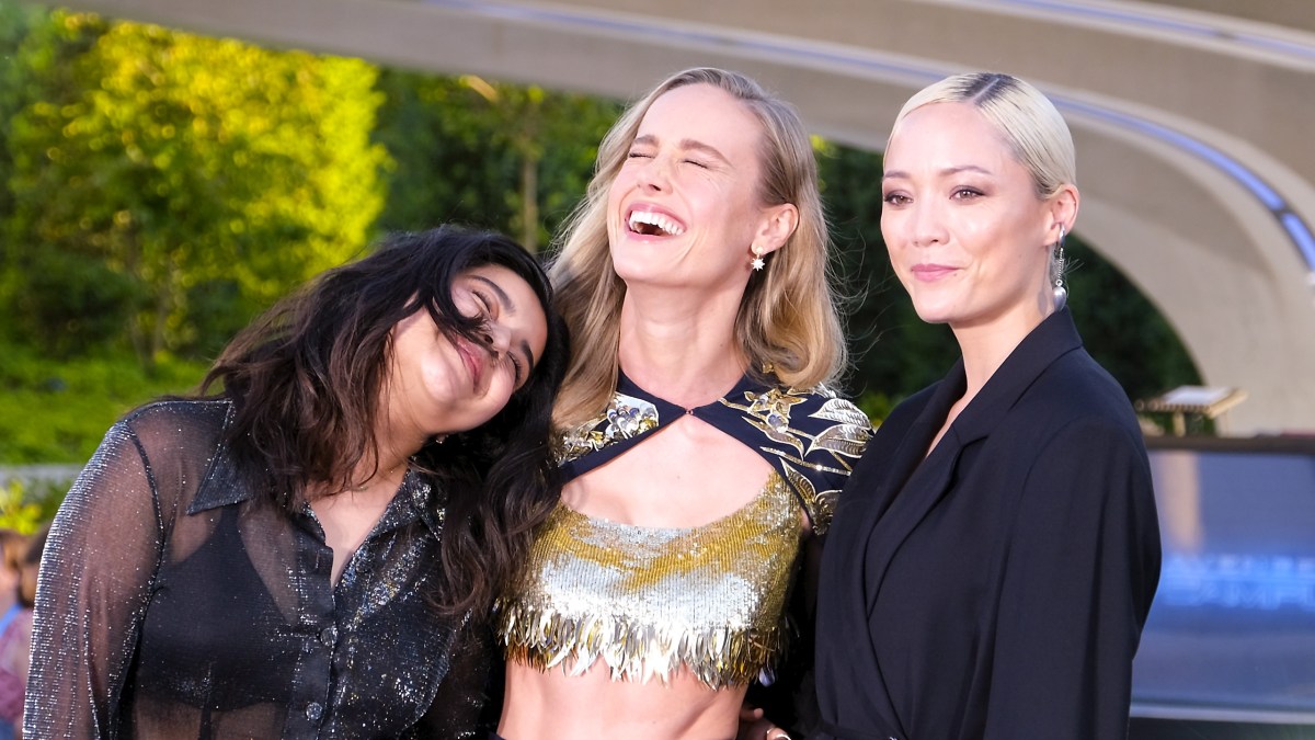 Brie Larson is pictured outdoors at a press event in a sparkly god cropped top and dramatic cape, laughing, with her head thrown back, flanked by Iman Vellani, who is also laughing, and Pom Klementieff, both wearing black