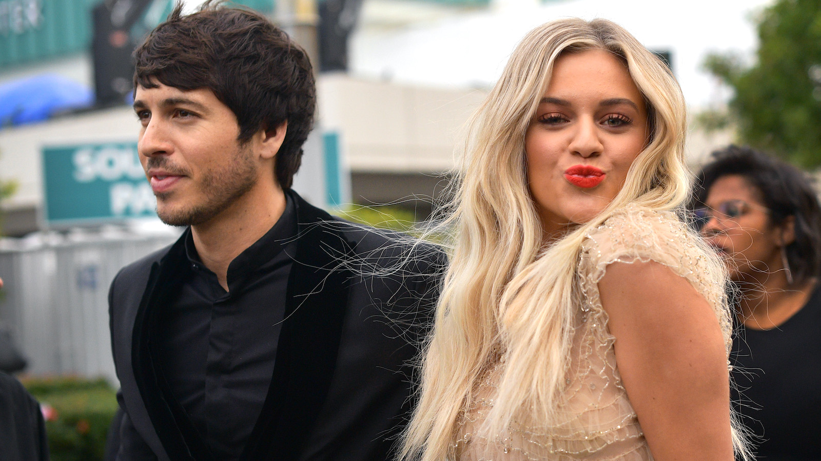 (L) Morgan Evans looking away from the camera in a dark suit and (R) Kelsea Ballerini blowing a kiss at the camera with red lipstick and a tan dress