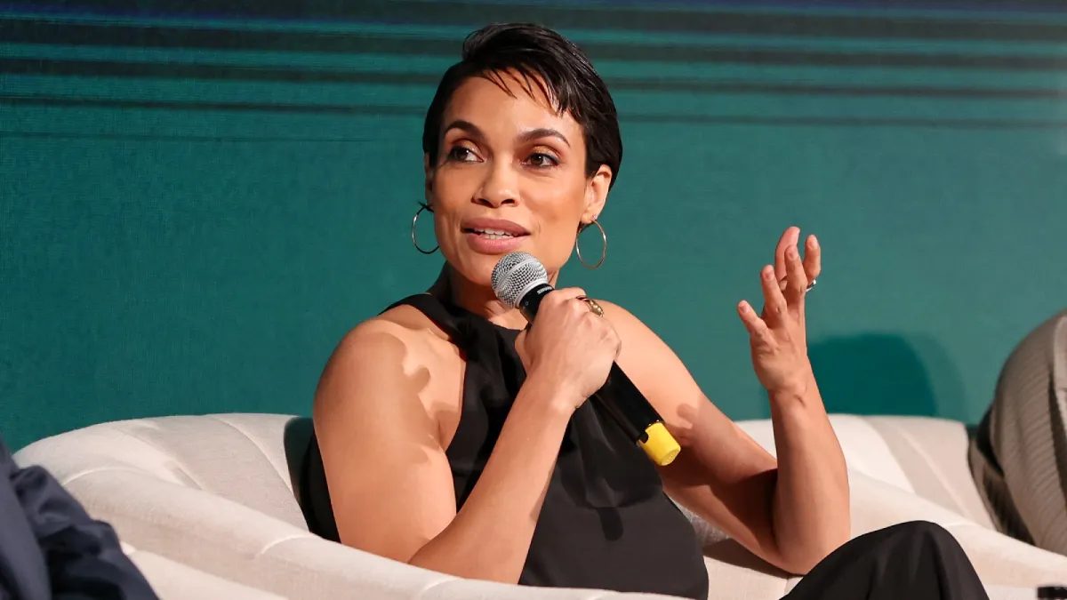 Rosario Dawson talks into a microphone while seated on a couch at a panel.