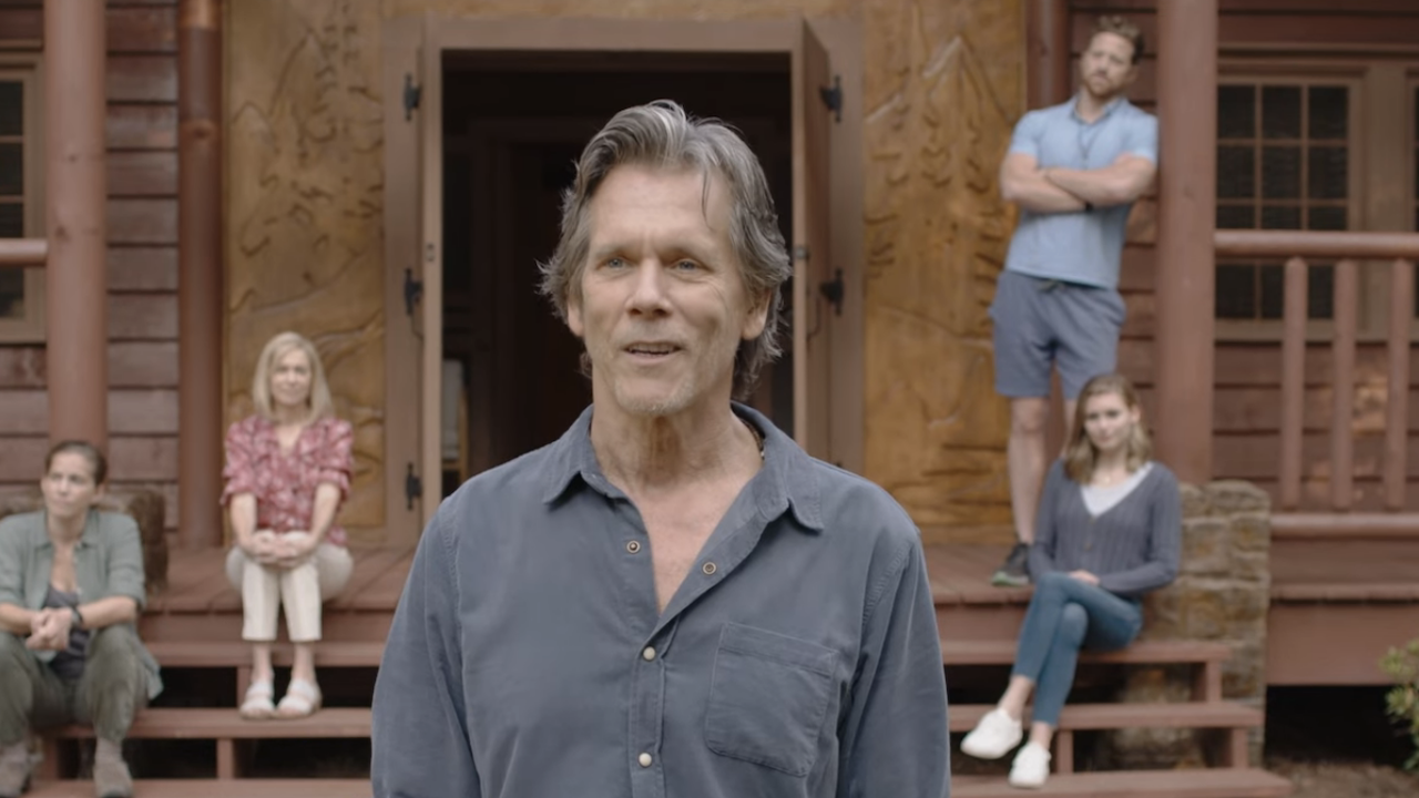 Kevin Bacon in character in They/Them, foregrounded and semi-smiling, in front of a wooden cabin