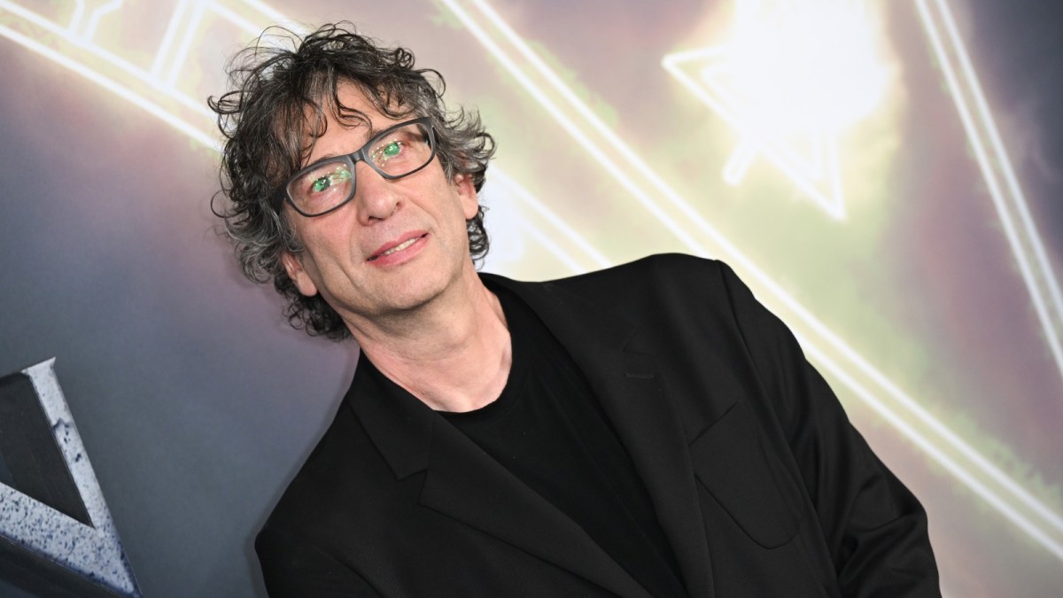 Neil Gaiman strikes a pose wearing a black blazer and glasses at a red carpet event.