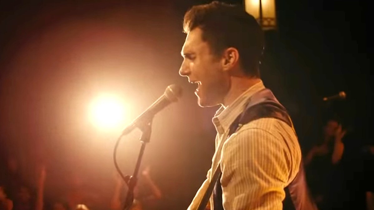 Profile shot of Adam Levine signing into a microphone and playing the guitar in the movie 'Begin Again'