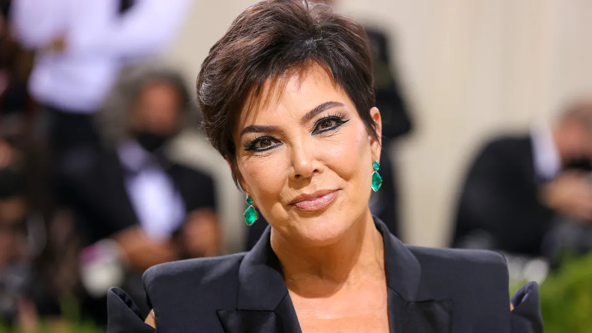 Kris Jenner pictured with black shoulder pads, emerald green earrings, her signature short air, and a closed smile while looking directly at the camera