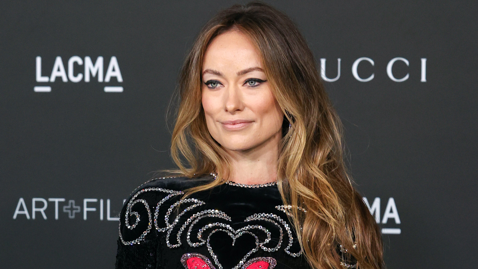 Olivia Wilde smiling in a black dress with wavy hair draped over one shoulder