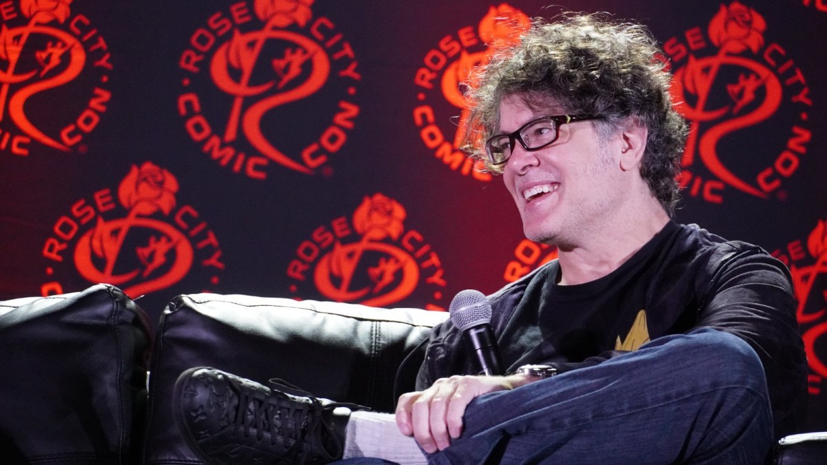Sean Schemmel holds a microphone while sitting on a couch on a stage against a red background.