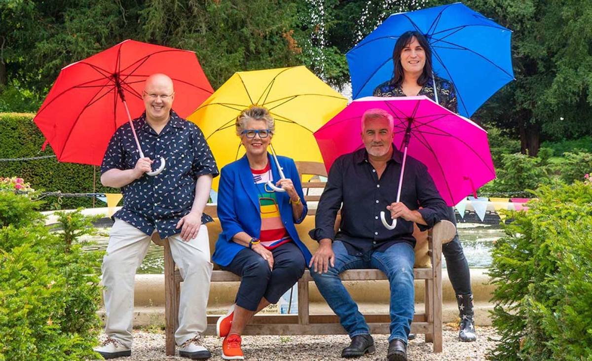 Sitting at a bench holding open colored umbrellas from left to right, Matt Lucas, Prue Leith, Paul Hollywood, and Noel Fielding from 'The Great British Baking Show'