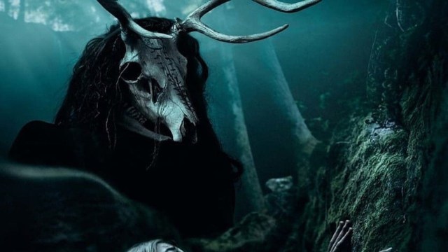 A man wearing a black robe and a deer skull against a murky blue blackground
