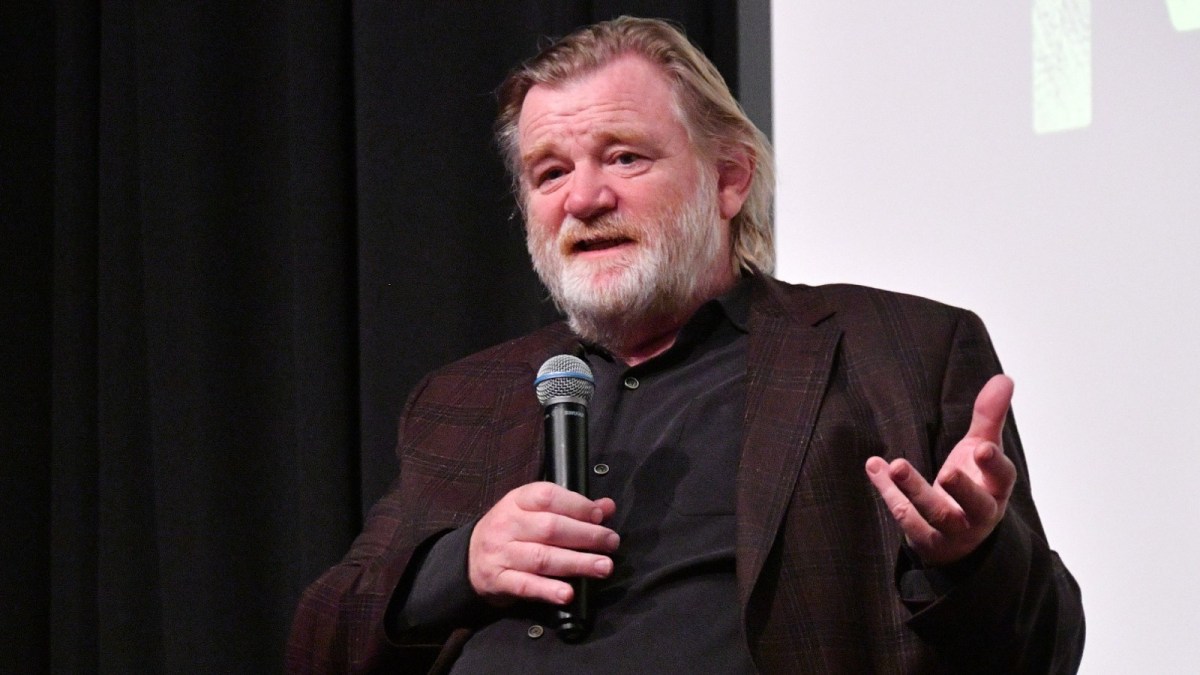 Brendan Gleeson speaks into a microphone at a stage during a press event.
