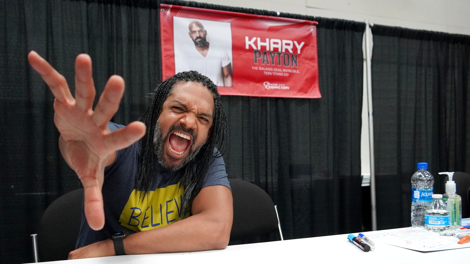 Khary Payton makes a dynamic pose into the camera while occupying a booth to greet fans at a comic con event.