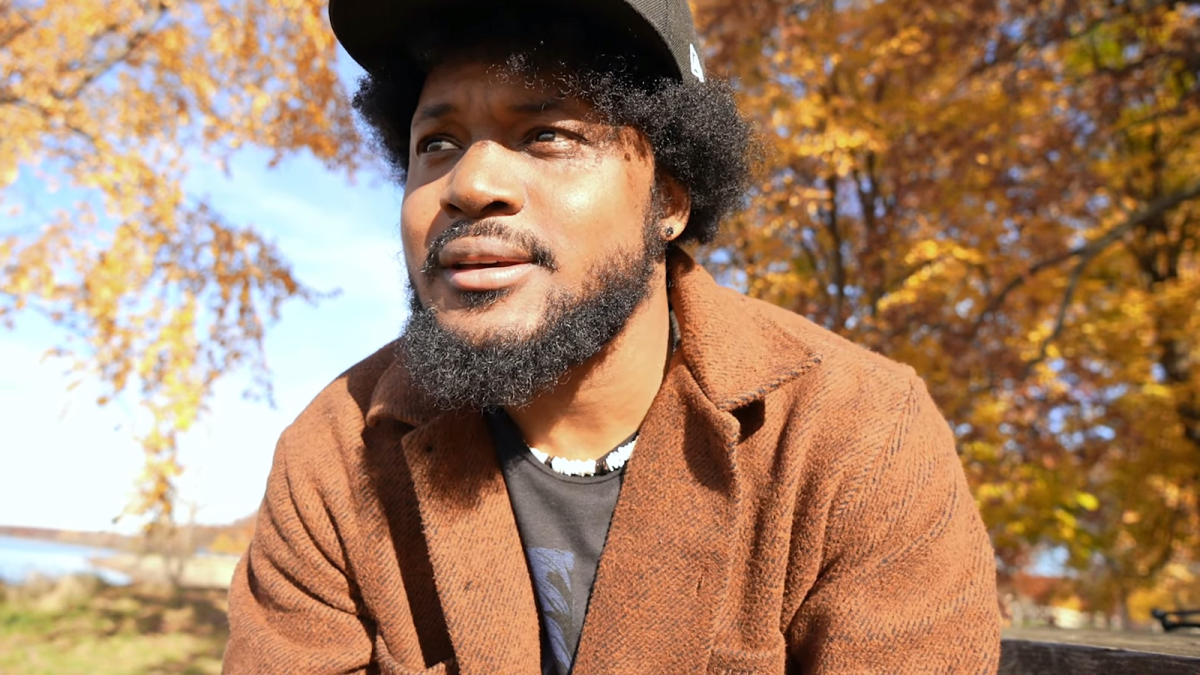 CoryxKenshin sitting on a bench at a park wearing a flat billed hat surrounded by orange leaves