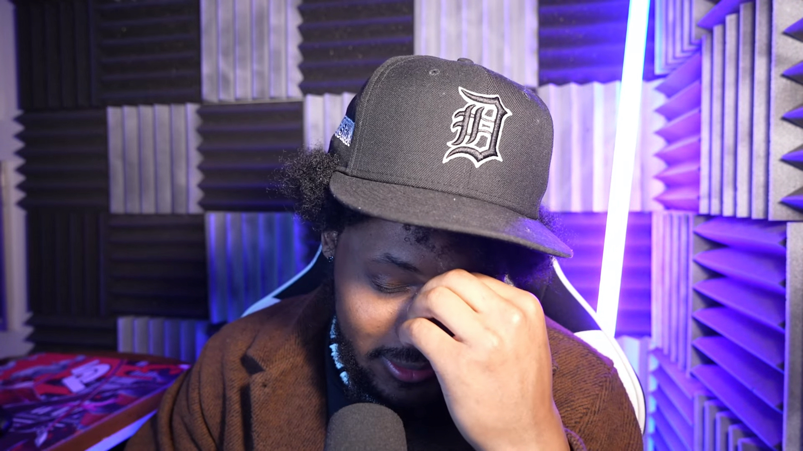 CoryxKenshin in a flat billed hat, pinching his nose, and looking down