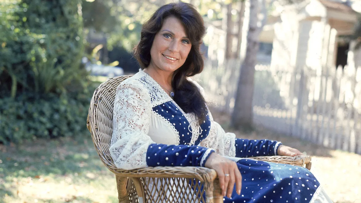 A young Loretta Lynn sits in a wicker basket chair and smiles at the camera