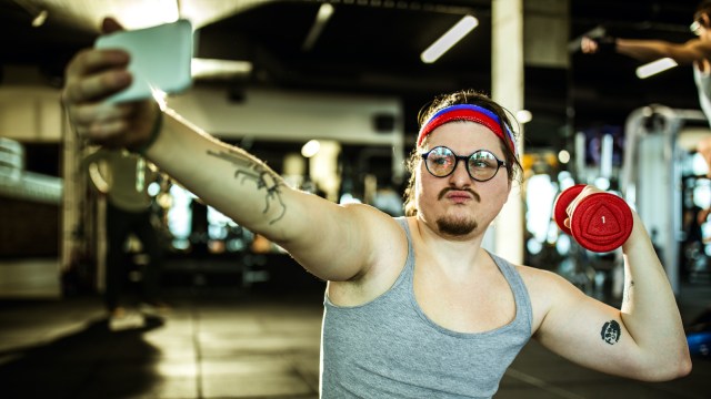 Nerdy athlete showing off while exercising with dumbbell in a gym and taking a selfie with smart phone.