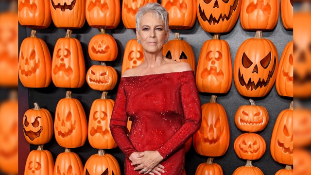 Jamie Lee Curtis poses against a backdrop of bright orange jack-o-lanterns while wearing a red sequin dress.