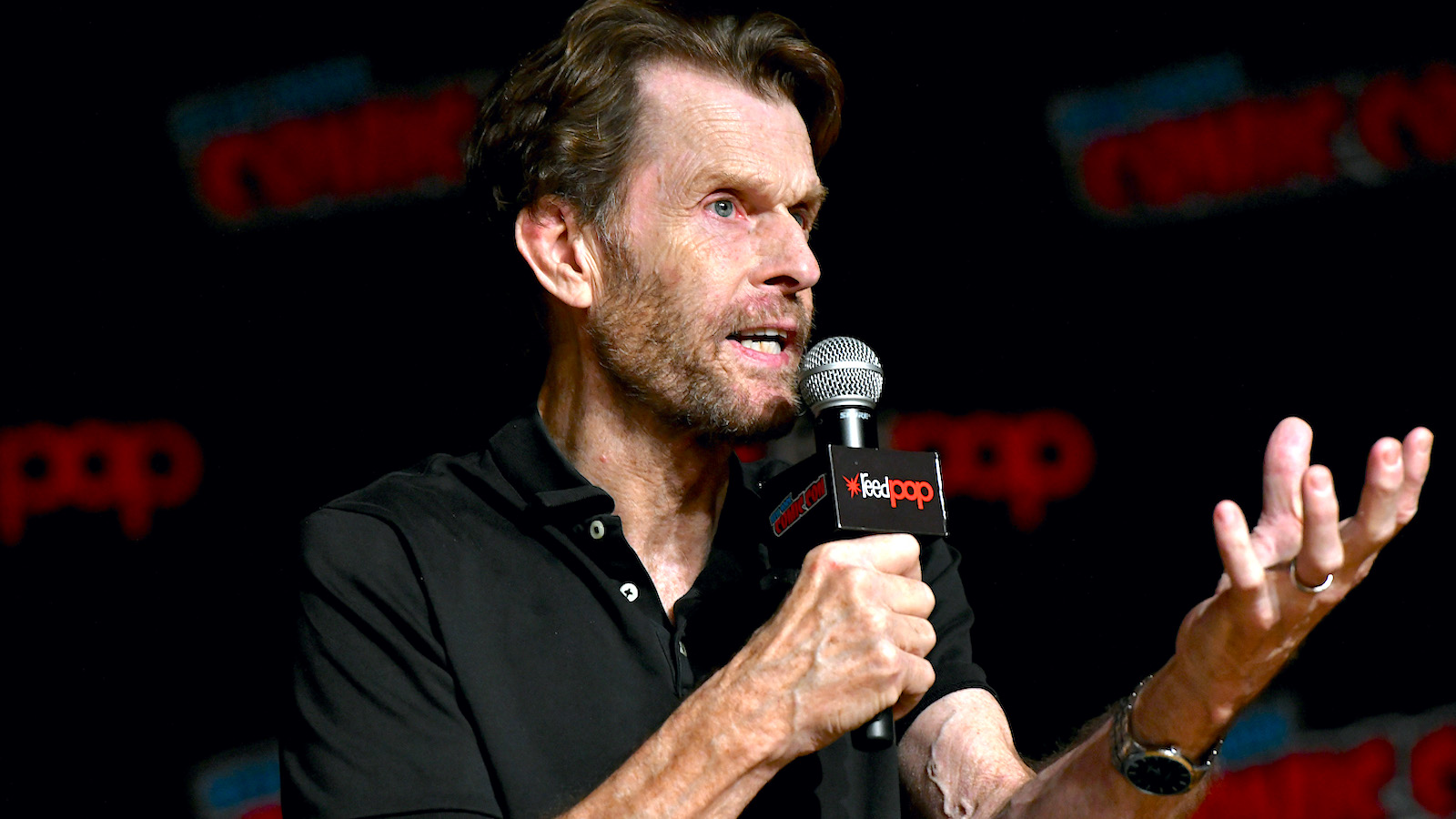Kevin Conroy speaks into the microphone wearing a black polo shirt