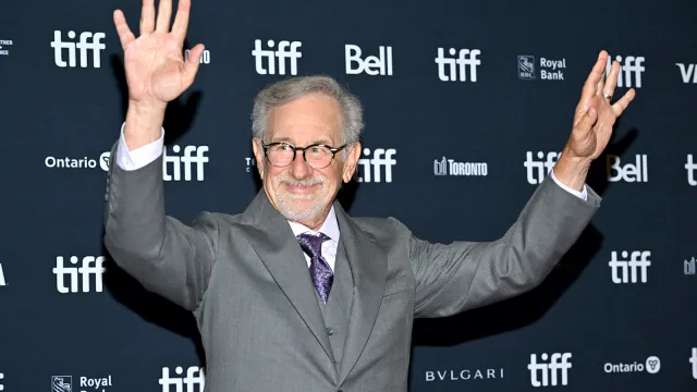 Steven Spielberg with his arms in the air wearing an all grey suit and purple tie on the red carpet.