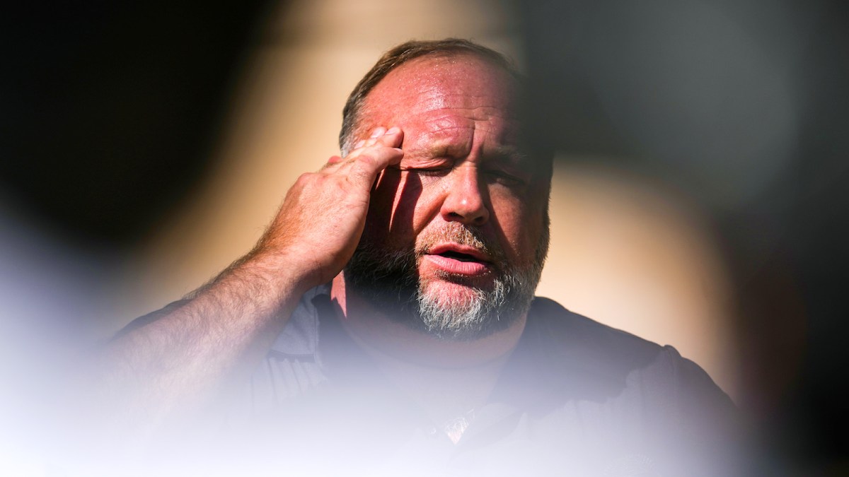 Alex Jones with one hand to his temple, eyes closed, looking defeated