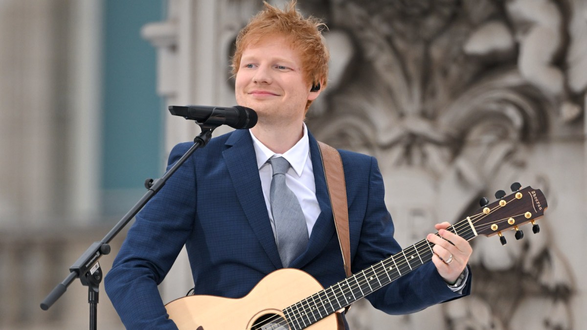 Ed Sheeran wearing a blue suit with an acoustic guitar strapped around his shoulder