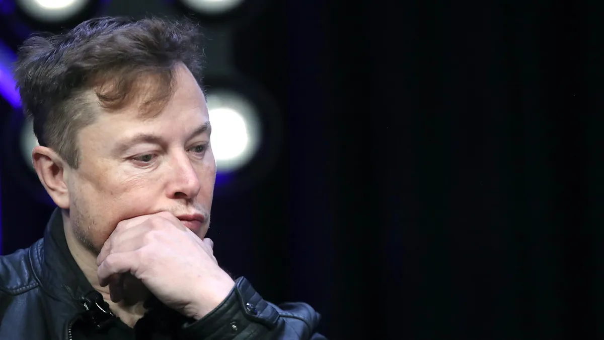 Elon Musk with his chin resting on hand, looking down.