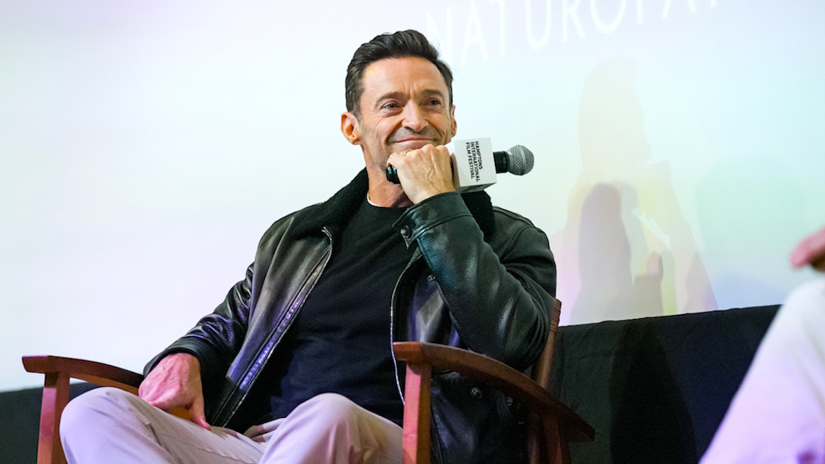 Hugh Jackman, dressed in a black shirt and black leather jacket, holding a microphone and resting his chin on his fist as he smiles at an interviewer off camera.