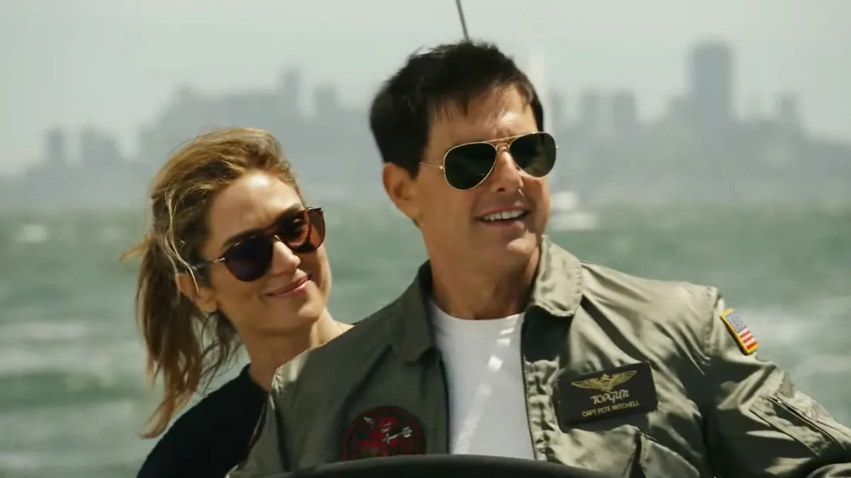 Tom Cruise as Maverick is smiling against a backdrop of ocean waves as he is steering a ship and wearing a Navy jacket and sunglasses. Jennifer Connelly's Penny is standing behind him, also in shades and her hair done up in a pony tail, and also smiling.