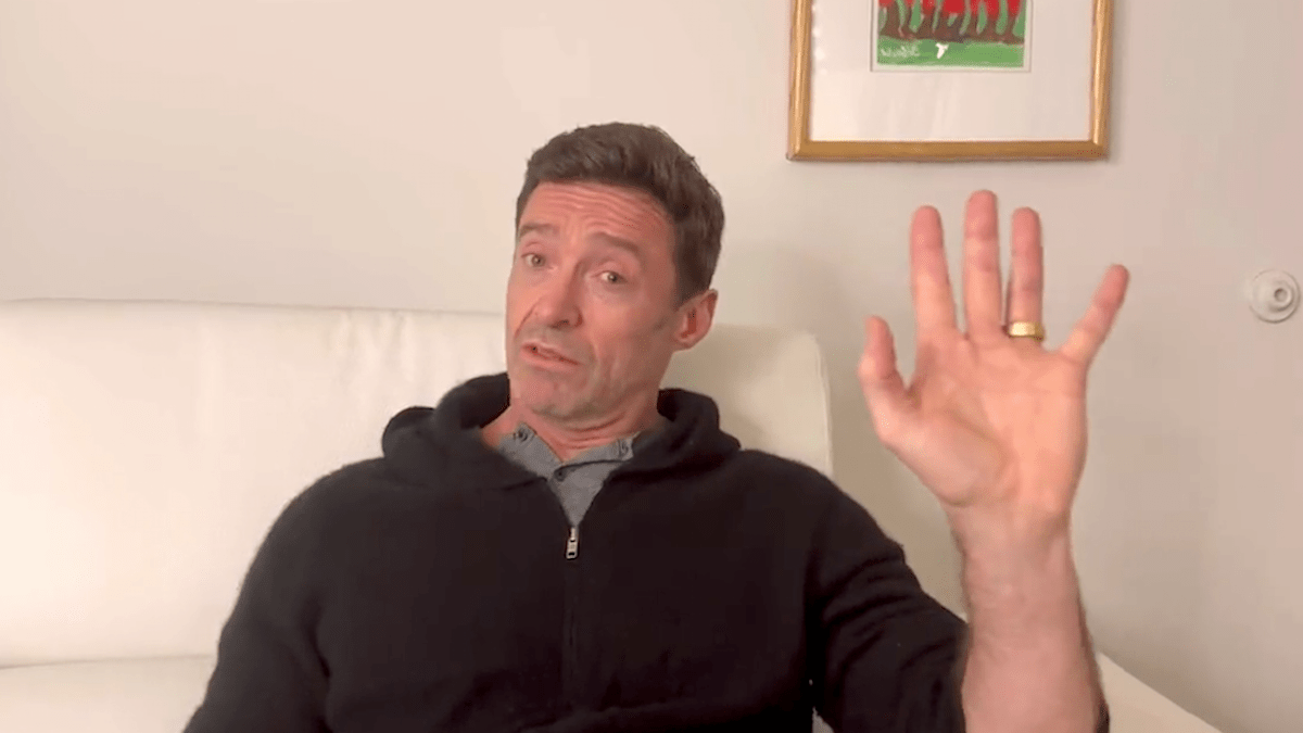 Hugh Jackman sitting on a couch wearing a black sweater, speaking to the camera