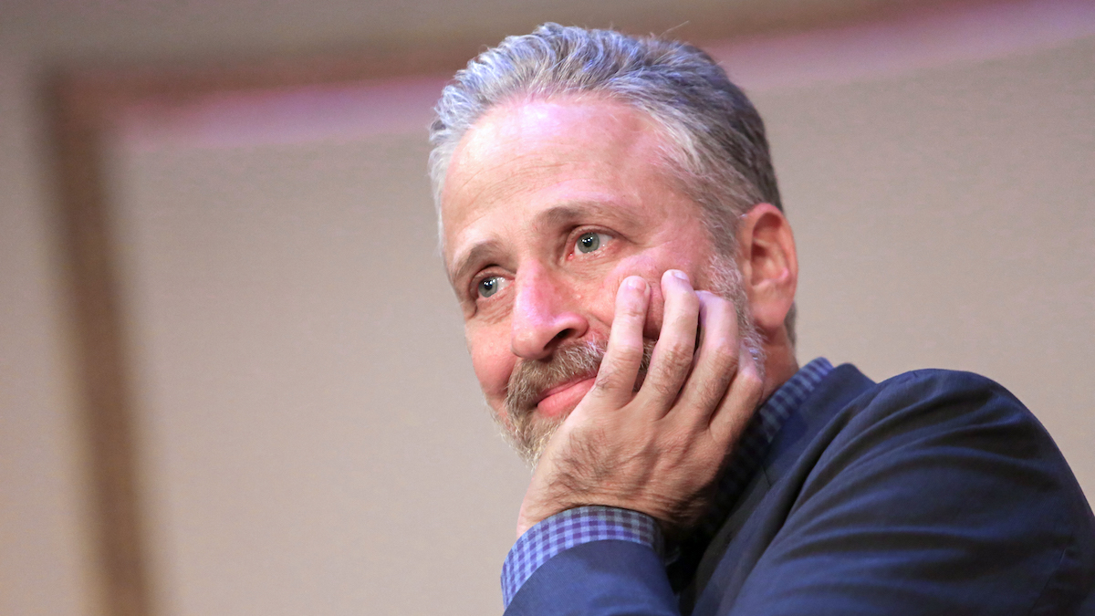 Jon Stewart rests his chin on his hand, smiling