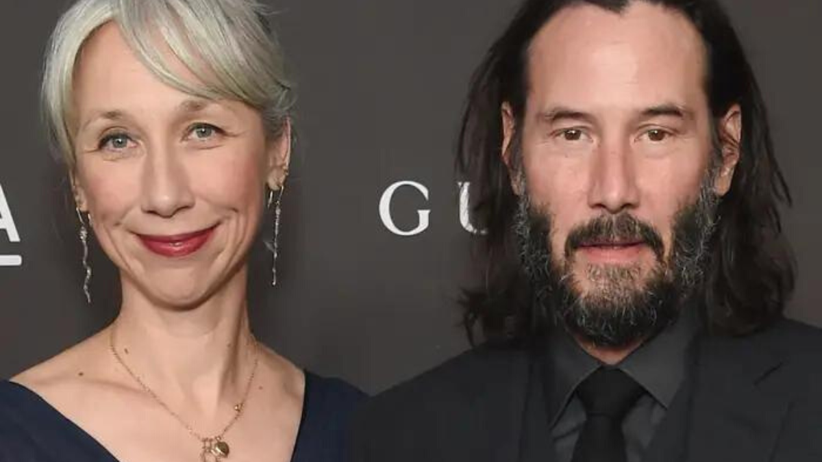 Alexandra Grant and Keanu Reeves posing next to each other for a photo on the red carpet, standing in front of a black backdrop. 