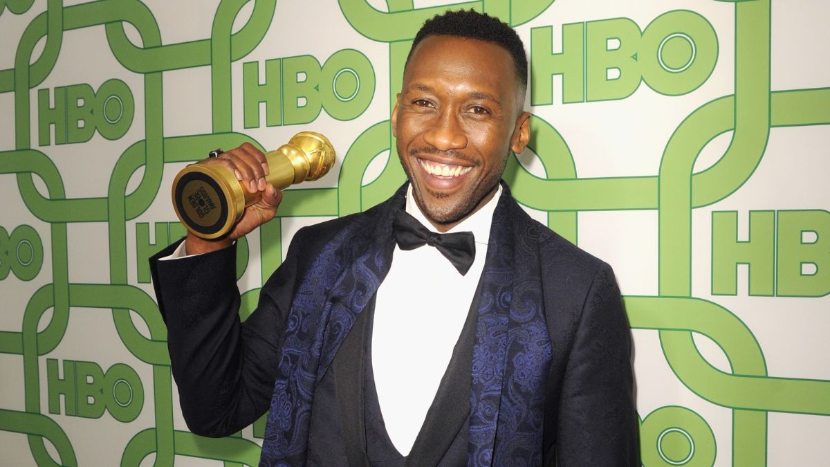 Mahershala Ali smiling in a suit while holding up a golden statuette