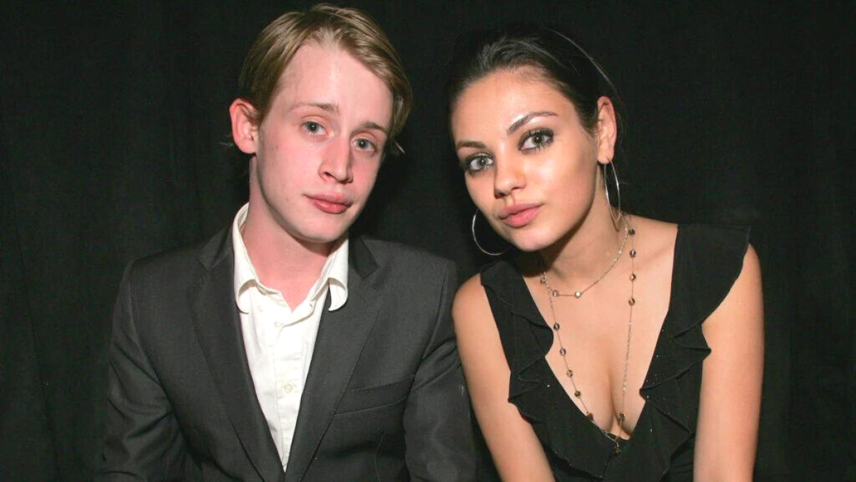 Mila Kunis and Macaulay Culkin, both wearing black, sit together and look at the camera. 