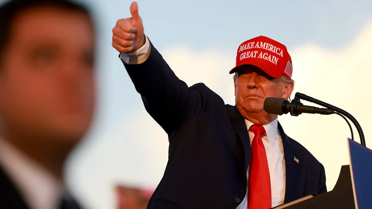 Donald Trump in a MAGA hat raising his arm and sticking out his thumb