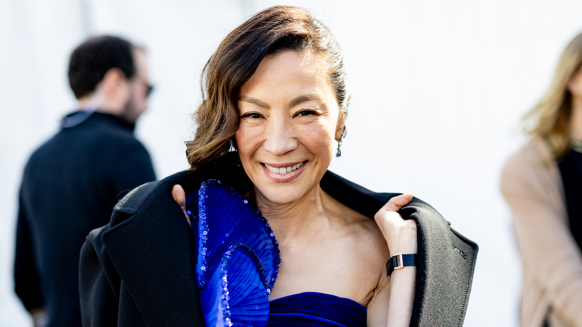 Michelle Yeoh smiling in a blue dress, her shoulders draped in a black coat