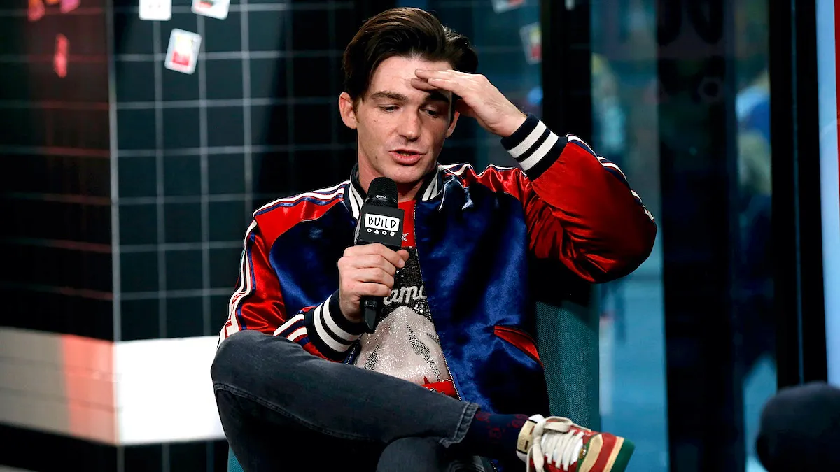 Drake Bell wearing a blue and red puffer jacket while sitting on stage holding a microphone and holding one hand to his forehead.