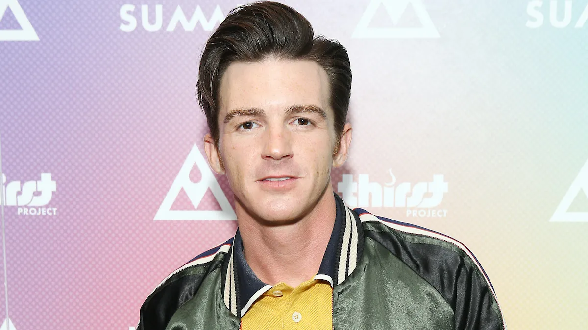 Drake Bell wearing a green puffer jacket and a yellow shirt, standing in front of a pale pink background