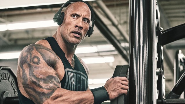 Dwayne "The Rock" Johnson is wearing headphones and a tank top at the gym.