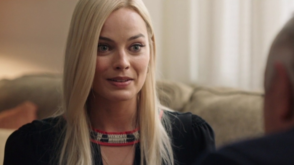 Margot Robbie is wearing black and sitting on a couch, while speaking to someone across from her. 